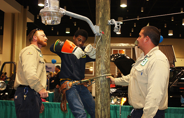 Two SRE linemen instructing a local 8th grade student how to climb a pole and wire an outdoor light.